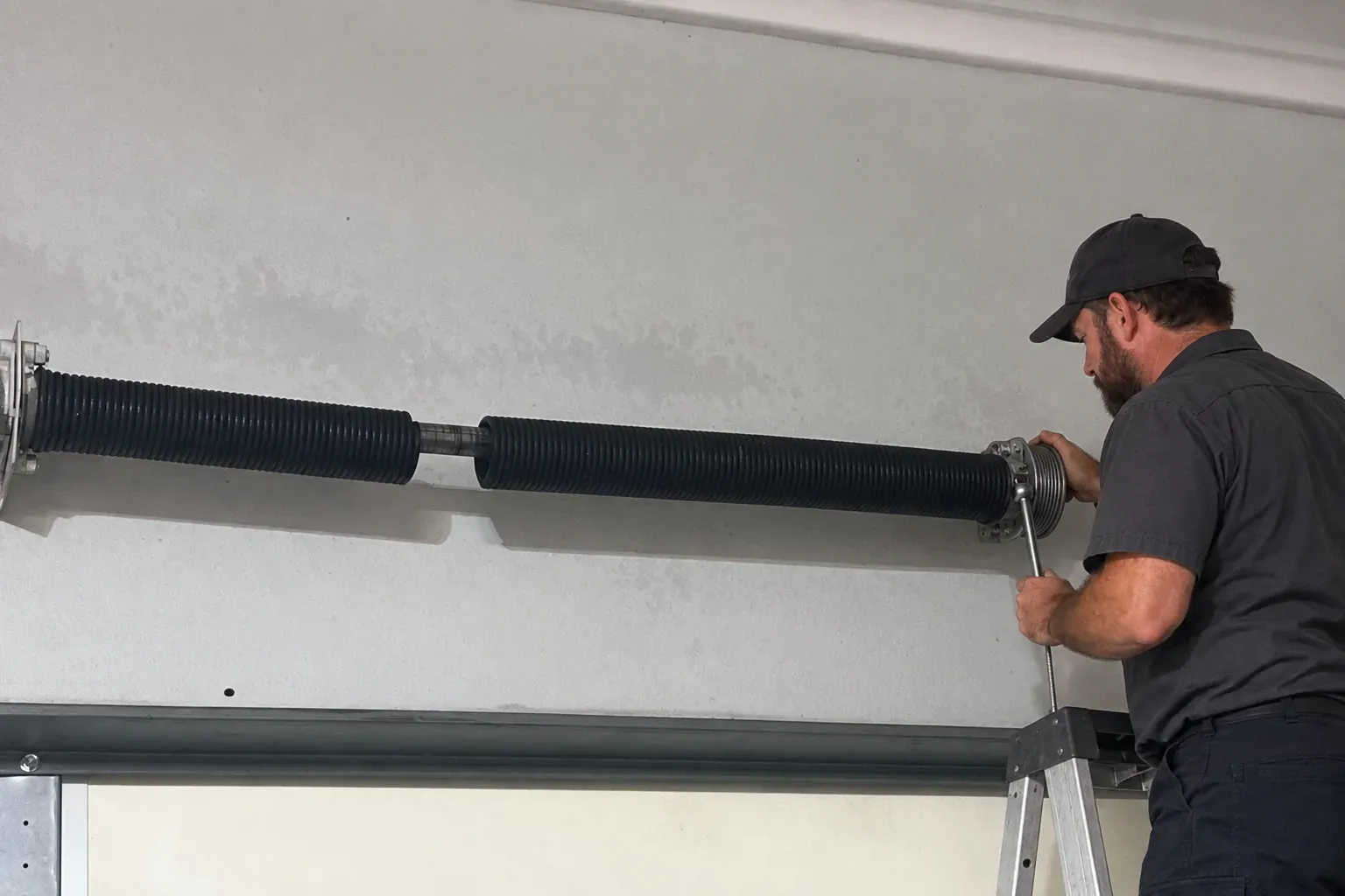 A technician on a ladder at the cable drum of a garage door, checking the lift cable