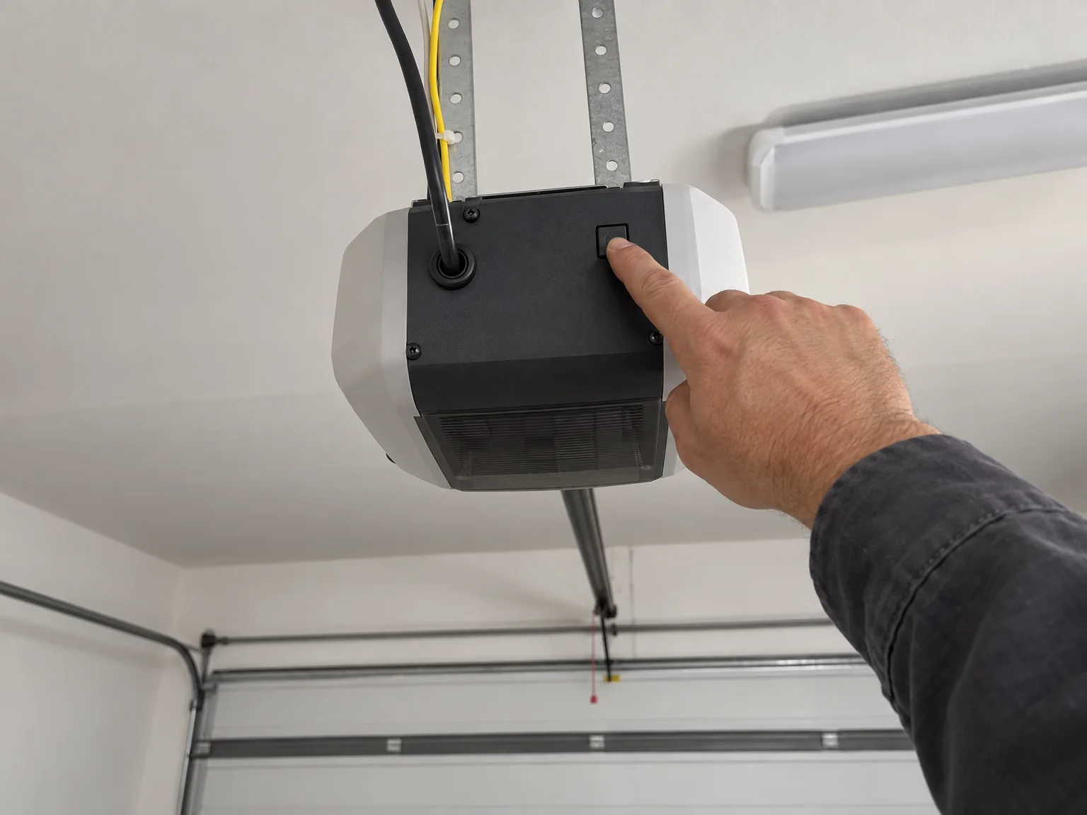 A technician’s finger on the small button at the back of a garage door opener on the ceiling
